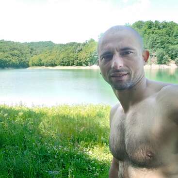 A shirtless man is taking a selfie outdoors beside a calm lake surrounded by lush green trees, under a bright and clear sky with natural sunlight.