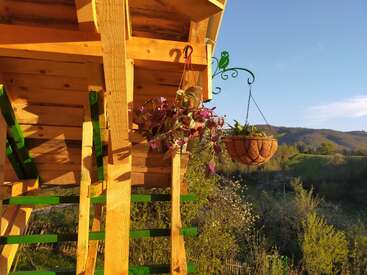A wooden terrace with hanging potted plants is shown in warm sunlight, overlooking green hills and trees under a clear blue sky. Peaceful rural scenery.