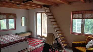 This cozy room features a bed, vibrant rugs, yellow armchair, exercise bike, wooden ceiling, staircase, large windows with blinds, and lots of natural light.
