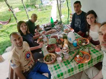 Six people are enjoying a meal together at a checkered table outdoors. The table is filled with food, drinks, and smiles, creating a warm atmosphere.