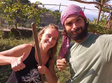 The image shows a man and woman posing for a photo in a garden, with the woman holding a long stick and giving a thumbs-up.