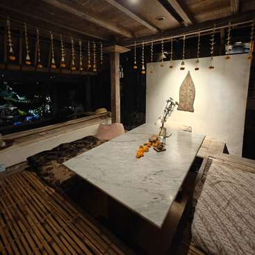 Un comedor acogedor y rústico con una mesa de mármol, suelo de bambú, cojines mullidos, guirnaldas de flores colgantes, una iluminación cálida y elementos decorativos minimalistas con toques naturales.