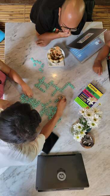 Three people play a word game at a marble table, surrounded by laptops, a dictionary, snacks, flowers, and a phone, creating a cozy, studious atmosphere.