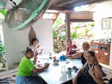 Five people are sharing a meal around a white table in a cozy, open-air setting. Everyone is smiling; one person makes a peace sign. Relaxed atmosphere.