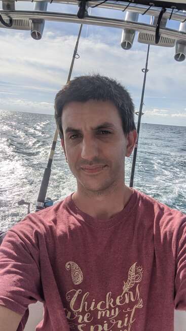 Ein Mann macht ein Selfie auf einem Boot und trägt ein kastanienbraunes Hemd mit Text. Im Hintergrund sind Angelruten und das Meer zu sehen, der Himmel ist teilweise bewölkt.