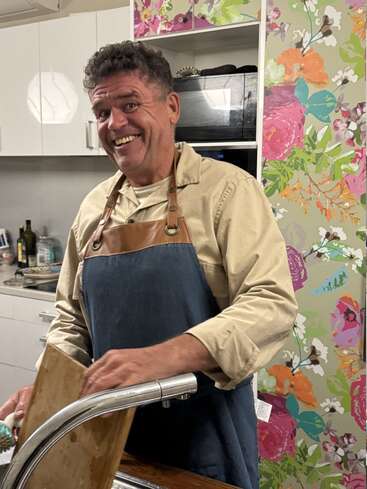 Un hombre sonriente con delantal azul lava los platos en un fregadero. El fondo muestra un colorido papel pintado de flores y armarios blancos. El ambiente es alegre.