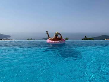 Une personne se détend sur une structure gonflable rose dans une piscine à débordement, un verre à la main, avec une vue imprenable sur l'océan et les montagnes sous un ciel dégagé.