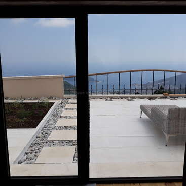 Par une porte vitrée, on aperçoit un patio moderne avec des dalles en pierre, un banc en osier et une vue pittoresque sur l'océan et la côte sous un ciel partiellement nuageux.