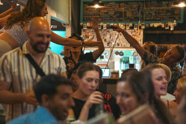 Une scène animée dans un bar où les gens boivent un verre, discutent et chantent au karaoké. L'ambiance est vibrante, énergique et animée par les rires et le mouvement.