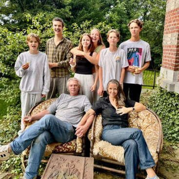 Um grupo de oito pessoas, provavelmente uma família, posa ao ar livre em sofás antigos, rodeadas por vegetação. Alguns estão em pé, outros sentados, sorridentes e relaxados.