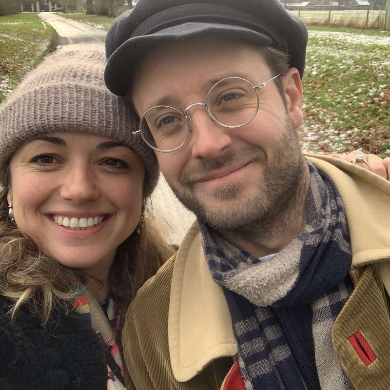 A imagem mostra uma selfie de um homem e uma mulher em pé do lado de fora, em uma área gramada com manchas de neve no chão, sorrindo para a câmera.