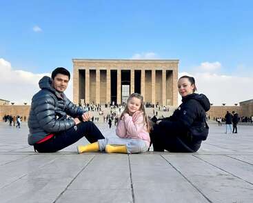 Une famille de trois personnes est assise sur une place pavée devant un grand monument, profitant d'une journée lumineuse avec un ciel bleu et des nuages épars.