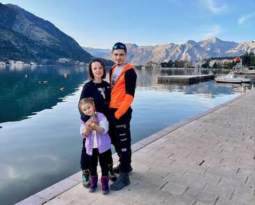 Une famille de trois personnes pose au bord d'un lac serein avec des montagnes en arrière-plan, profitant d'une journée ensoleillée sur une promenade au bord de l'eau avec des bateaux à proximité.
