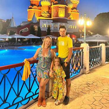 Une famille de trois personnes pose au bord d'une piscine éclairée la nuit, avec en arrière-plan une architecture colorée et ornée ressemblant au design russe, magnifiquement illuminée. Ils sourient.