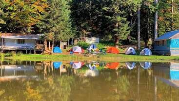 Barracas coloridas e duas cabanas estão instaladas ao longo de uma tranquila margem do lago, cercadas pela floresta. Seus reflexos brilham lindamente na água calma e tranquila.
