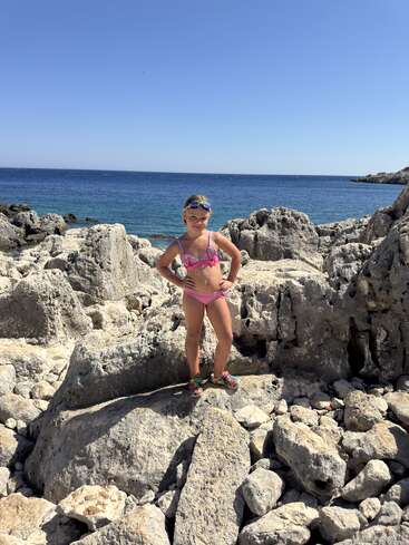 Uma jovem com um maiô rosa e óculos de proteção está confiante em um terreno rochoso à beira-mar, sob um céu azul claro, aproveitando um dia ensolarado.