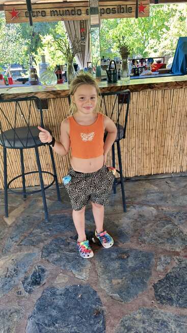 Uma criança está confiante em um ambiente interno, usando um top laranja brilhante e shorts de leopardo, com sapatos coloridos. Atrás deles há uma área de bar rústica decorada com plantas.