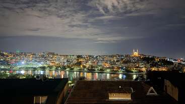 Uma vista noturna de tirar o fôlego de uma cidade com prédios iluminados, uma mesquita bem iluminada, um rio refletindo as luzes da cidade e um céu nublado e dramático acima de você.