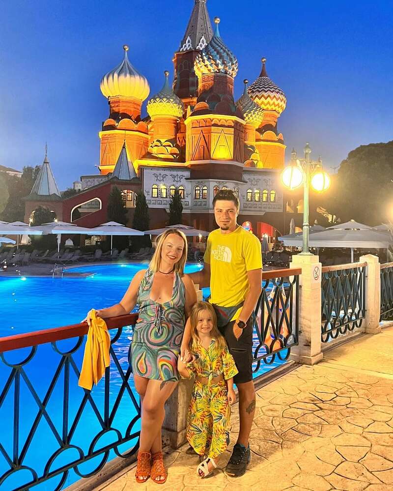 Uma família de três pessoas posa em uma piscina bem iluminada à noite, com uma arquitetura colorida e ornamentada que lembra o design russo, iluminada lindamente ao fundo. Eles estão sorrindo.