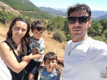 A family of four poses outdoors on a sunny day, surrounded by lush green hills, pine trees, and mountains in the scenic background. They are smiling.