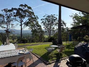 Auf der friedlichen Terrasse stehen Tische und Stühle zum Essen bereit, umgeben von grünem Gras, hohen Bäumen, fernen Bergen und einem klaren blauen Himmel im Hintergrund.