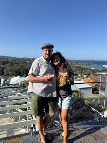 Ein lächelndes Paar steht auf einem Balkon und genießt einen Drink. Hinter ihnen liegt eine malerische Küstenlandschaft mit blauem Himmel, Meer, Grünflächen und Wohnhäusern.