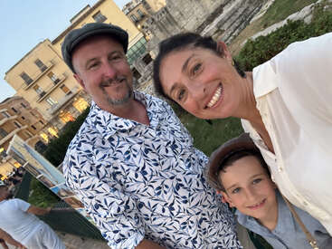 Una sonriente familia de tres miembros se toma un selfie al aire libre frente a antiguas ruinas y viejos edificios, disfrutando juntos de una agradable tarde de vacaciones.