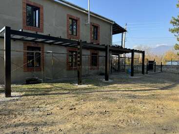 A two-story house under construction features metal scaffolding and a black steel frame structure at the front, set in a yard with dirt and sparse grass.