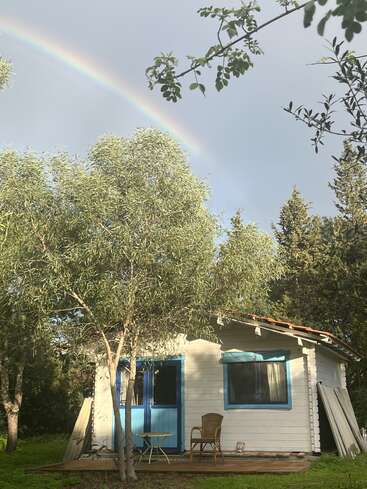 Eine kleine weiße Hütte mit blauen Verzierungen liegt friedlich zwischen grünen Bäumen. Ein Regenbogen spannt sich darüber, während ein Stuhl und ein Tisch auf der Veranda stehen.
