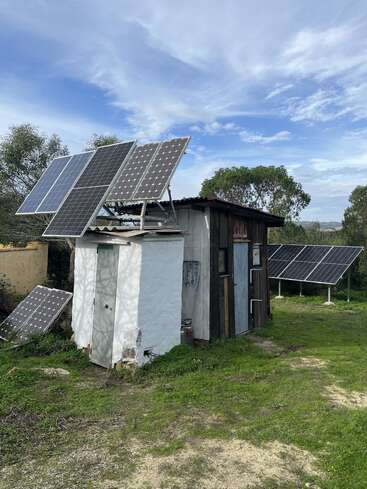 Das Bild zeigt ein kleines Gebäude mit mehreren Solarzellen auf dem Dach und in der Nähe des Bodens, umgeben von Grün und unter einem teilweise bewölkten blauen Himmel.