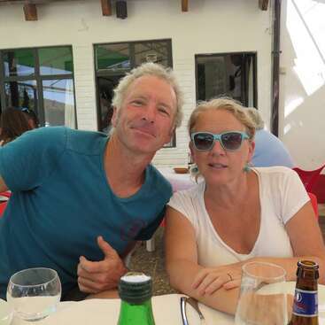 A imagem mostra um homem e uma mulher sentados em uma mesa, com o homem vestindo uma camisa azul e a mulher com uma blusa branca, ambos aparentando ter 50 ou 60 anos.