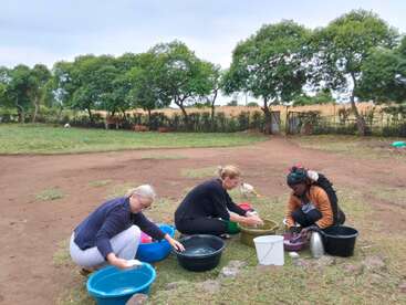 Trois femmes sont assises par terre, en plein air, et lavent du linge dans des bassines en plastique. Derrière elles, un paysage rural avec des arbres verts, des vaches et un champ.