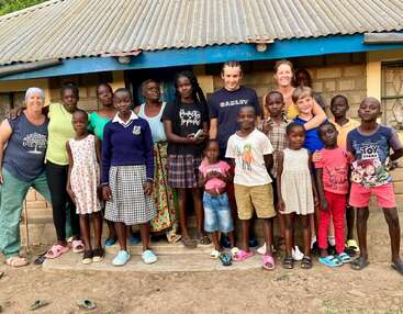 Un groupe diversifié d'enfants et d'adultes sourient ensemble devant une petite maison, illustrant l'unité, la joie et la communauté dans un environnement rural.