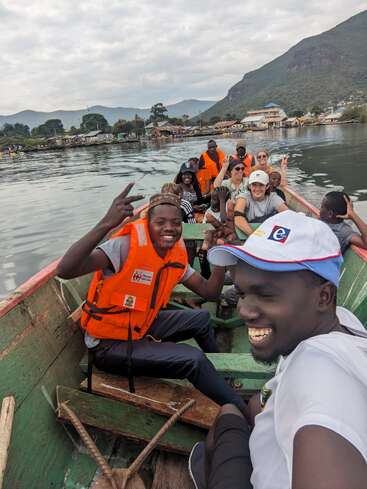 A group of cheerful people wearing life jackets are riding in a wooden boat on a calm lake, with mountains and a small village in the background.