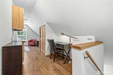 Cette image montre une chambre mansardée lumineuse et étroite, avec des plafonds en pente. On y voit un bureau, une chaise, une armoire, une petite penderie et un fauteuil rouge confortable près d'une fenêtre.