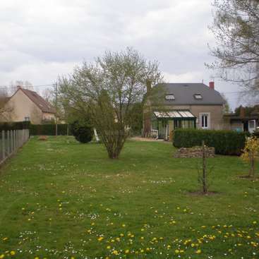 Das Bild zeigt eine ruhige Vorstadtszene mit einem Haus, einem Baum und einem Blumenfeld vor einem bewölkten Himmel.