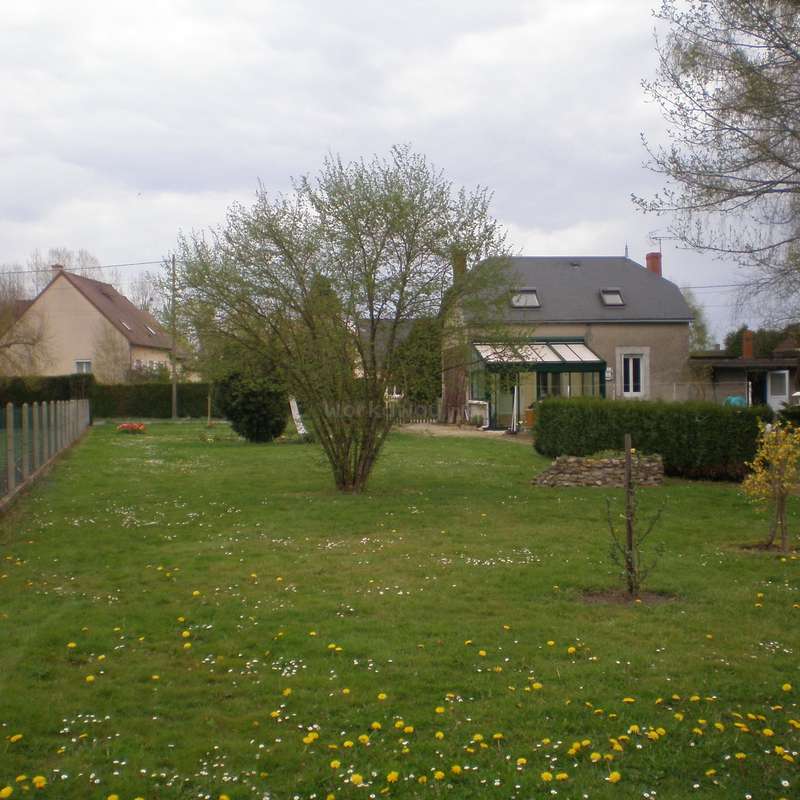Das Bild zeigt eine ruhige Vorstadtszene mit einem Haus, einem Baum und einem Blumenfeld vor einem bewölkten Himmel.