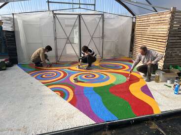 Three people are painting a vibrant, colorful mural with swirling, spiral patterns and wavy stripes on the floor inside a partially enclosed, greenhouse-like structure.