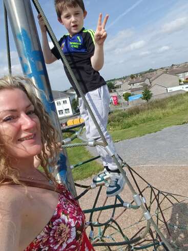 Une femme et un jeune garçon sourient sur une structure de corde à grimper dans une aire de jeux. Le garçon fait un signe de paix par une journée ensoleillée.