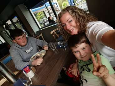 Une femme et deux garçons sont assis à la table d'un restaurant. La femme et le plus jeune garçon sourient pour un selfie, tandis que le plus âgé mange un dessert.