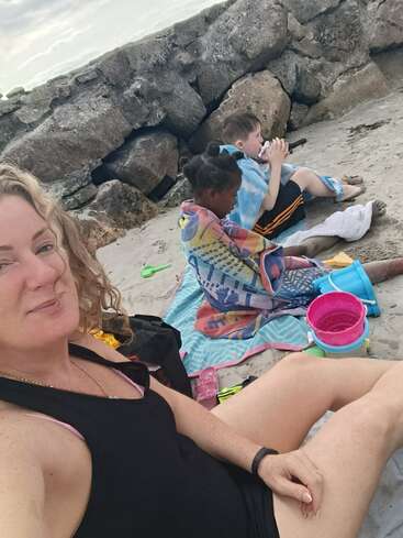 Une femme prend un selfie à la plage avec deux enfants. Ils sont assis sur des serviettes près de rochers, enveloppés dans des serviettes colorées, et s'amusent avec des jouets de sable et des seaux.