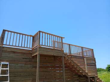 Une plate-forme d'observation en bois, dotée de balustrades et d'escaliers, se détache sous un ciel bleu limpide. Une échelle métallique est appuyée contre la structure, avec de l'herbe verte tout près.