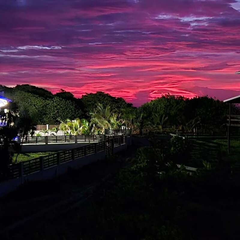 A imagem mostra uma paisagem tropical serena ao pôr do sol, com uma ponte, edifícios e vegetação exuberante, em um céu rosa e roxo vibrante.