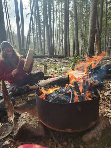 Una persona sonriente está sentada en un bosque, tocando un tambor junto a una hoguera de colores vivos. Entre los objetos que la rodean hay una pluma, un termo, un tarro y una exuberante vegetación.