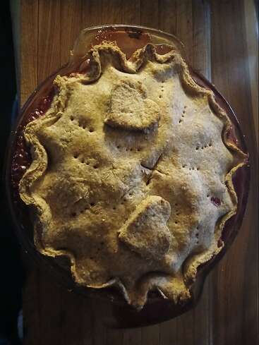 Una tarta casera de estilo rústico con una corteza dorada, bordes ondulados y adornos en forma de corazón. El relleno burbujea ligeramente y descansa sobre una superficie de madera. Acogedora y tentadora.