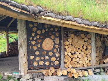 Esta imagen muestra una leñera rústica con un tejado de césped vivo. La estructura está construida con leña y troncos apilados, lo que le confiere un aspecto natural y rústico en un entorno rural.