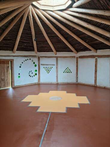 Se trata de una sala circular con vigas de madera que parten de una claraboya central, suelos en tonos tierra, un elemento central geométrico de color amarillo y detalles decorativos con botellas de cristal en las paredes.