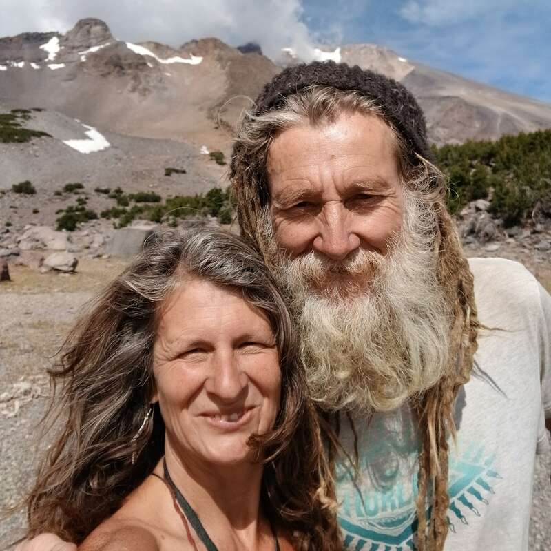 Una pareja sonriente con el pelo largo y barba posa al aire libre, rodeada de montañas rocosas, manchas de nieve, vegetación y un cielo nublado y luminoso. Parecen felices.