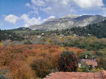 Paisaje con árboles otoñales, montañas con laderas rocosas, casas dispersas, un cielo azul brillante con nubes y tejados anaranjados en primer plano.