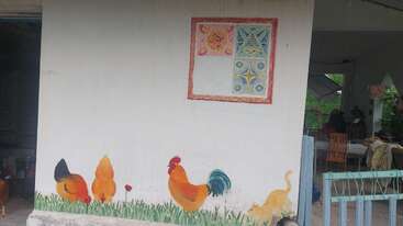 Una pared blanca presenta coloridos cuadros de dos gallinas, un gallo y un gato en la hierba. Encima, un motivo geométrico y floral enmarcado decora la pared.